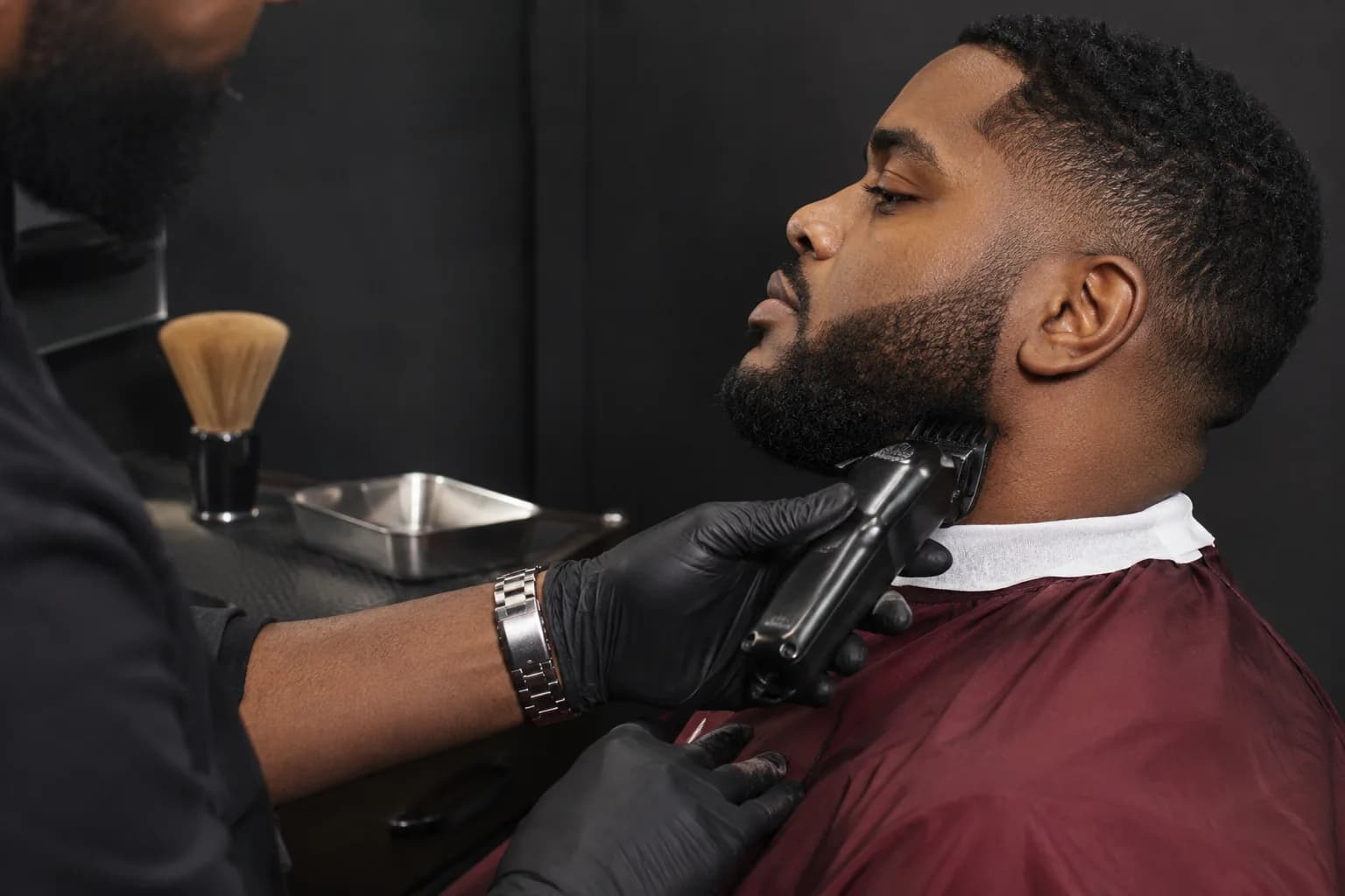 Adult barber using a guarded trimmer below an adult man's jaw in a calm close side view