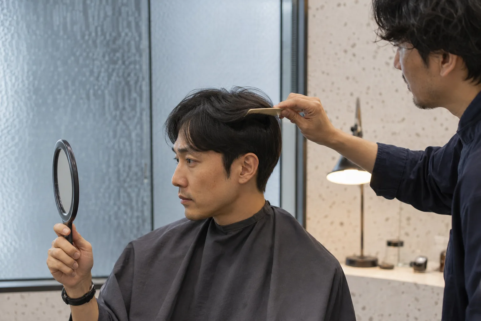 Adult barber lifting a section of an adult client's hair while he checks the side shape in a handheld mirror
