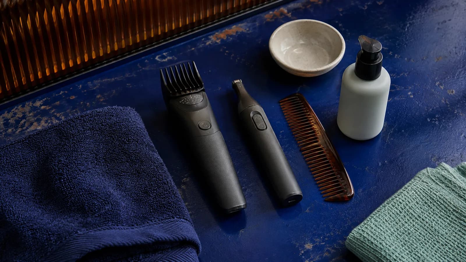 Guarded beard trimmer, detail trimmer, comb, towel, bowl and moisturizer arranged on a blue grooming surface
