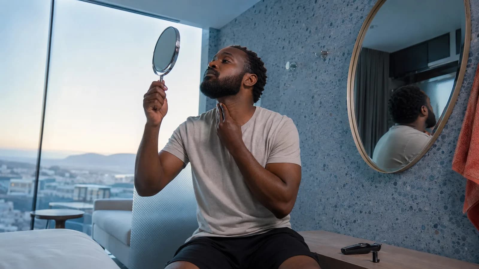 Adult man with a medium beard checking the underside neckline with a hand mirror in a cool blue hotel dressing room