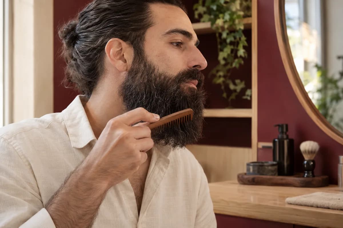 Hombre adulto con barba larga peina suavemente las puntas junto a un mueble de madera clara y pared burdeos