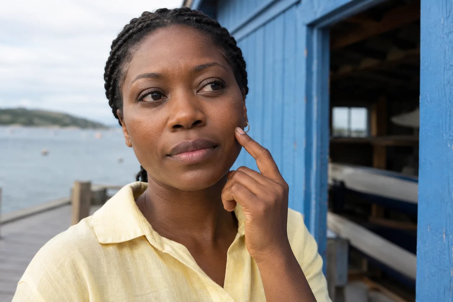 Femme adulte en chemise jaune pâle indiquant légèrement le haut de sa joue devant un hangar à bateaux bleu