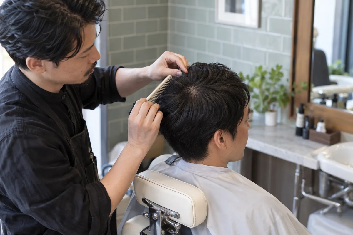 Coiffeur séparant doucement au peigne les cheveux au sommet de la tête d'un client adulte dans un salon ivoire et sauge