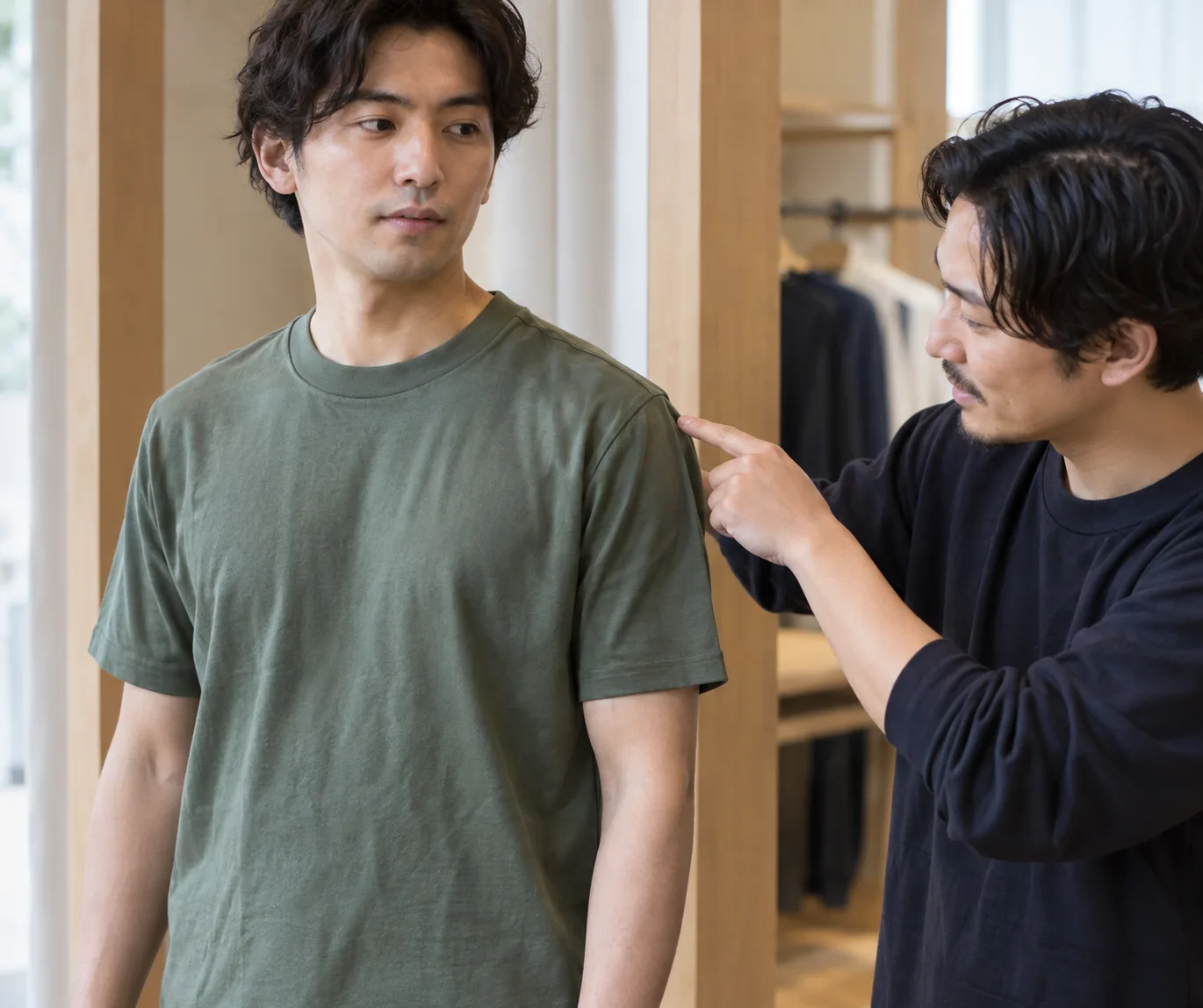 Adult man checks the shoulder seam of a plain crew-neck shirt with a friend in a fitting area