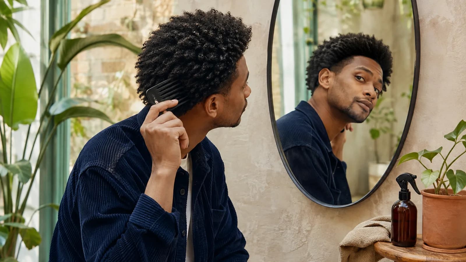 Adult Black man gently checking the side shape of medium coily hair with a wide-tooth comb in a plant-filled room