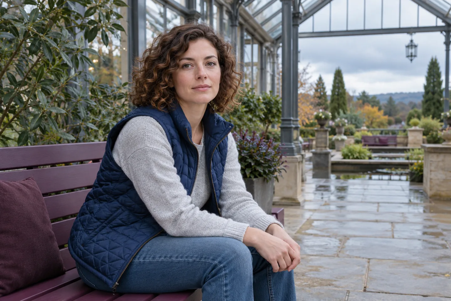 Femme adulte en gilet matelassé bleu assise calmement sur un banc prune sous une verrière près d’une cour arborée