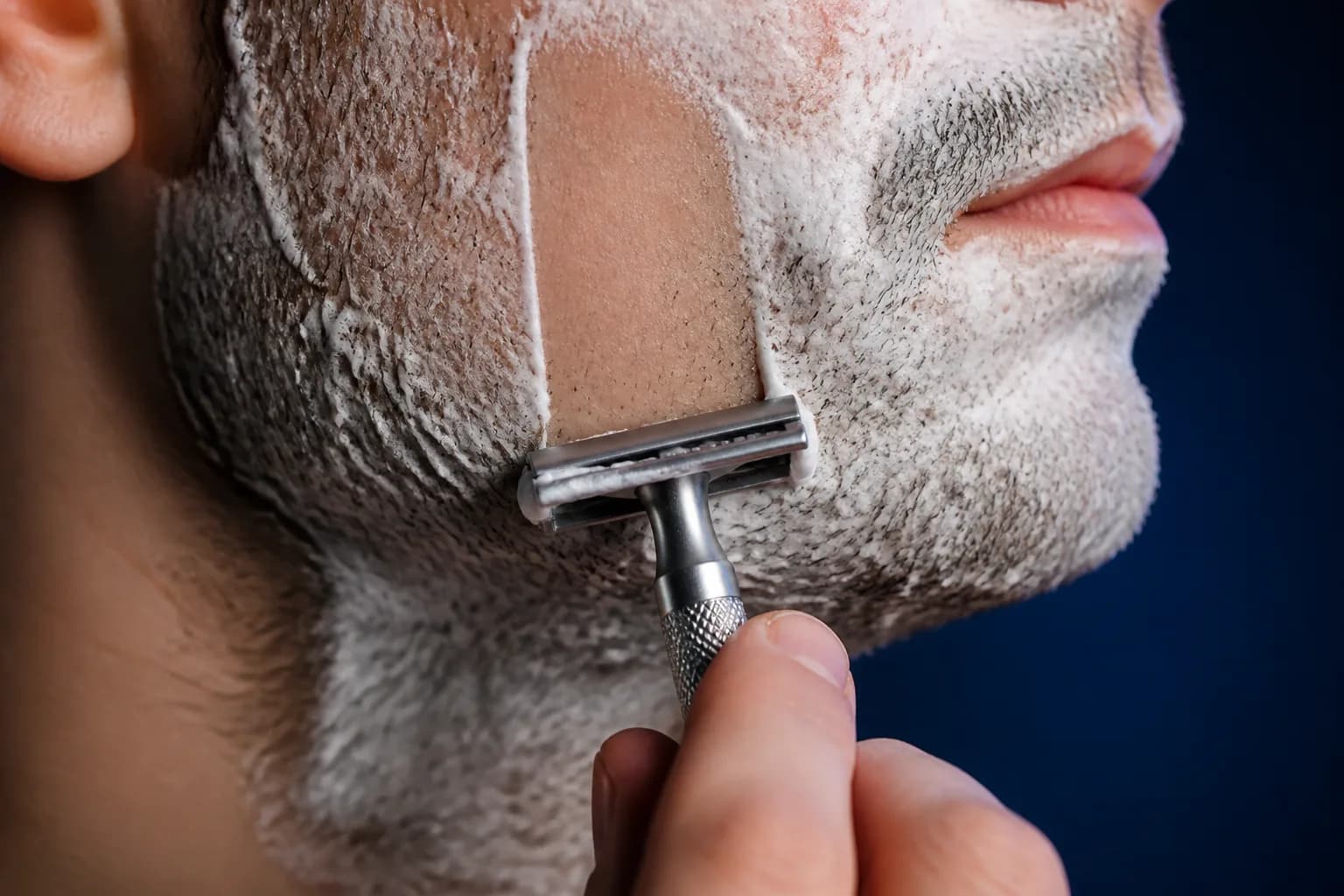 Close view of a safety razor making one short light pass through shaving cream along an adult man's jaw