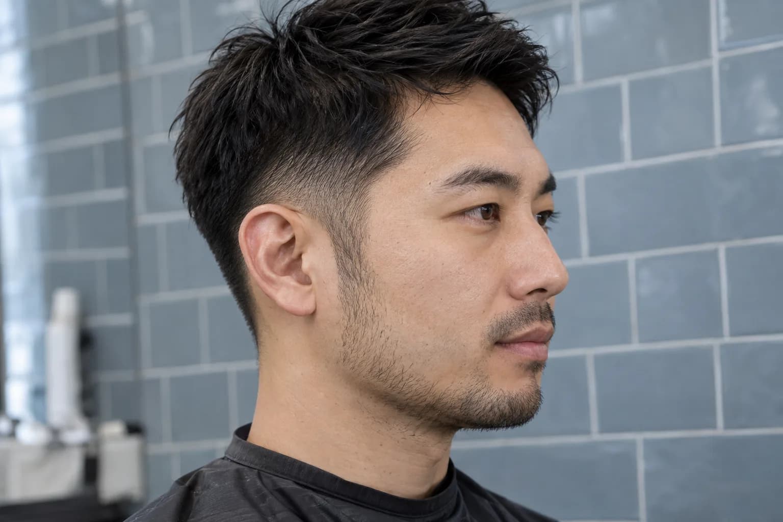 Adult man with short straight dark hair shows a natural sideburn transition into light stubble beside a grey-blue tiled wall